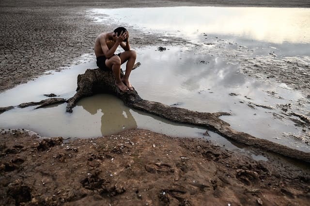 foto mostra menino sentado em um tronco em cima do que foi um rio um dia, foto alusiva à falta d'água, seca na amazônia, aquecimento global