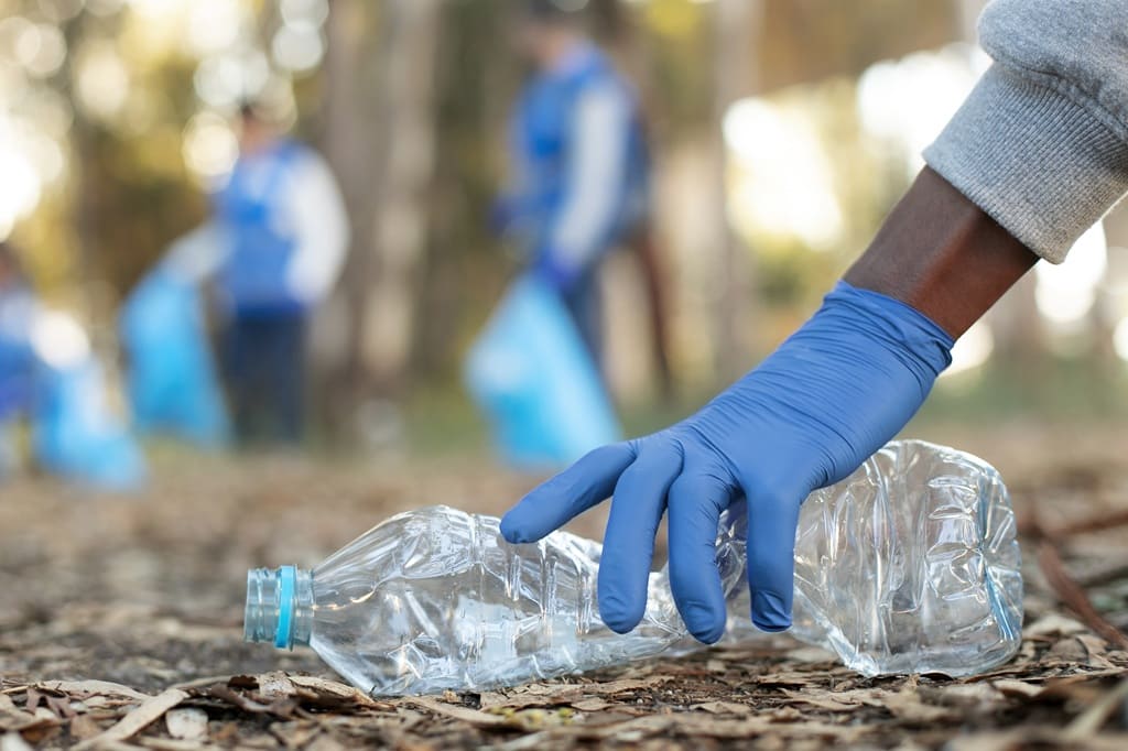 foto de mão de catador pegando garrafa plástica em parque arborizado, remete a matéria do dia mundial da reciclagem